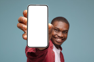 Fototapeta na wymiar Mobile advertisement. Overjoyed black man pointing at cellphone with empty white screen on blue studio background, mockup for app or website. Cellphone display template