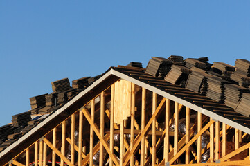 Obraz premium Close-up of wooden roof truss framing with stacked roofing tiles staged for installation, illustrating early-phase construction activity in residential single-family housing development projects