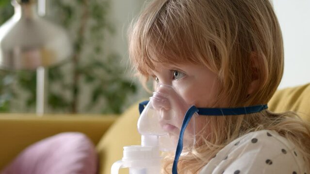 child girl sits at home on couch and makes inhalation using mask. beautiful curly toddler cures allergy or other respiratory disease. Doctor appointment for home health procedures.