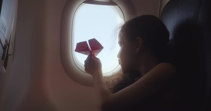 Child's hand with small paper plane against the background of airplane window. Child looking porthole. Child sitting by aircraft window and playing with little paper plane, during flight on airplane