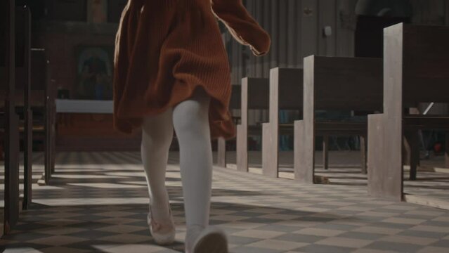 Cropped shot of unrecognizable little girl in pretty dress and white tights running along central walkway in church with sun rays coming from rose windows