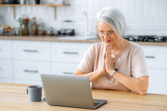 Satisfied middle aged caucasian gray haired woman with glasses, sitting at home in the kitchen at the table, looks at the laptop screen, wants something, worries, holds palms together, Generative AI