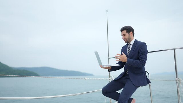 Caucasian businessman working and video conference on laptop computer with internet for corporate business during travel on luxury private catamaran boat yacht sailing in the ocean on summer vacation.