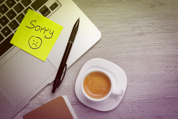 Sticky note with word Sorry and drawn sad face attached to laptop on table. Workplace with cup coffee, flat lay