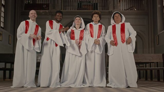 Five multiethnic church choir members in long white gowns clapping their hands and singing standing in row in old Catholic Church
