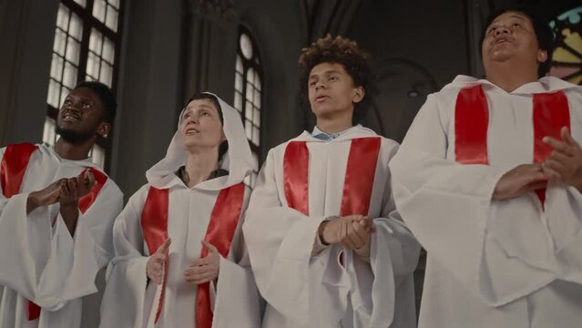 Low angle medium shot of delighted multiethnic church choir wearing long white gowns clapping and singing together standing together in old Catholic Church