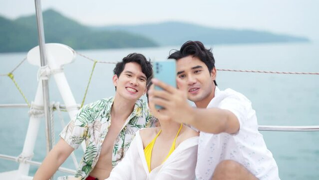 Asian man and woman friends enjoy outdoor lifestyle travel ocean on sailing luxury private catamaran boat yacht and using mobile phone taking selfie together on summer tropical holiday vacation.