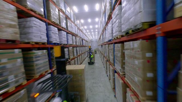 FPV drone flight between multi-storey shelves and over forklifts moving freight in frozen food storage facility warehouse