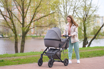 Fototapeta na wymiar Young beautiful smiling mother pushing stroller with newborn baby walking in spring park. Parenthood, healthy lifestyle concept 