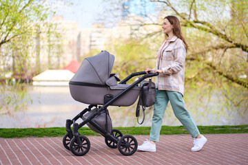 Portrait of beautiful smiling young mother pushing stroller with little baby walking in spring park. Motherhood, healthy lifestyle concept