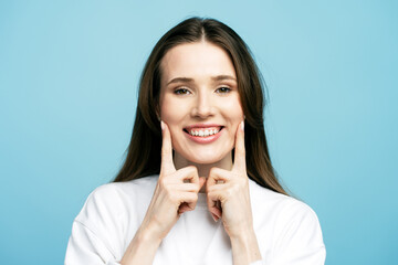 Closeup portrait of happy beautiful young woman with toothy smile pointing with fingers on his mouth looking at camera isolated on blue background. Health care, clinic,  dental treatment concept