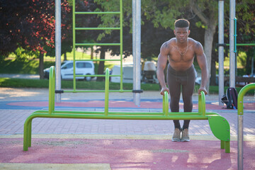 Young fit shirtless black man doing push-ups on parallel bars in a calisthenics park outdoors on...