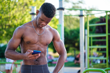 Portrait of a young fit shirtless black man using the smartphone in a calisthenics park outdoors....