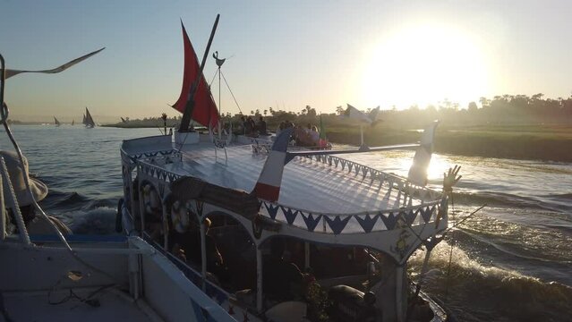 Slow Motion of Multiple egyption Local felucca tour boat on Nile river fully loaded with Tourists heading to Banana Island from Luxor, Egypt at sunset golden Hour passing by scenic fields on the sides