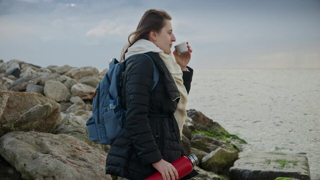 Woman with long hair, standing on the rocks and watching the winter waves while drinking hot tea. A great addition to travel or holiday-themed projects