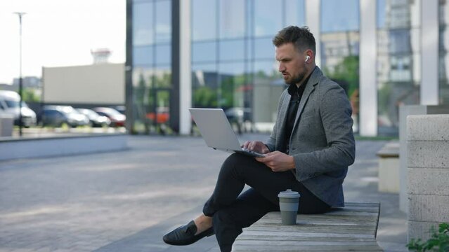 Young Businessman Sitting on the Bench and Working on the LapTop Wearing Earphones. It Manager Distance Working near Office Centre. Handsome Boss Online. Man Using his Laptop While Sitting Outdoors