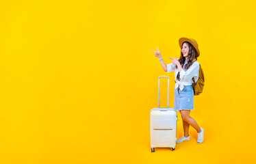 Pretty Asian woman passenger in trendy fashion is pointing while carrying her luggage bag in happiness for travel and summer vacation isolated on yellow background with copy space.