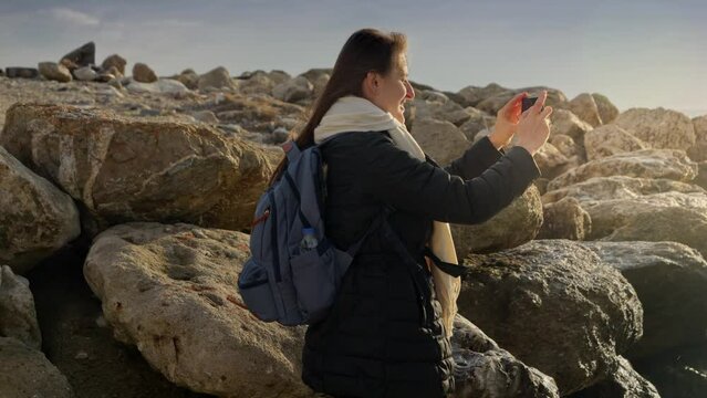 Young woman capturing the beauty of the winter sea by taking pictures with her smartphone while standing on the rocks. Ideal for vacation and travel