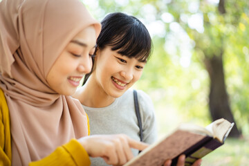 Happy beautiful Asian females Muslim student and friends reading a textbook together in a park. Diversity in ethnic and religious concept.