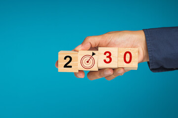 Hand businesspeople holding wooden cubes with the letters 2030 and dartboard against a blue background - obrazy, fototapety, plakaty