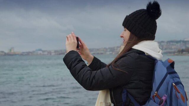 Young woman capturing the beauty of a stormy winter day at the beach with her smartphone, the waves crashing against the rocks behind her. Travel, adventure, and exploration