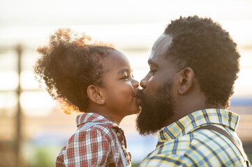 Happy african black parents dad father with daughter child on piggyback teasing fun in garden greenhouse. Black daughter kiss cheek dad and neck riding in vegetable greenhouse garden in light sunset