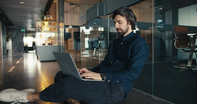 Successful Office Worker Working Comfortably on Laptop. Office worker sits on the floor near a glass wall and work on a laptop Generative AI