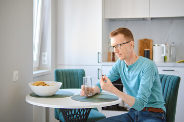 Middle aged man in the kitchen table and holding a pill and a glass of water in hands