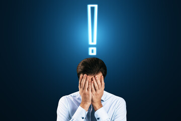 Worried young businessman covering face with hands. Glowing exclamation mark hologram above head on blue background. Stress, anxiety and confusion concept.