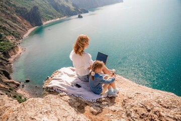 Freelance woman with her daughter working on a laptop by the sea, typing on the keyboard, enjoying the beautiful view, highlighting the idea of remote work.