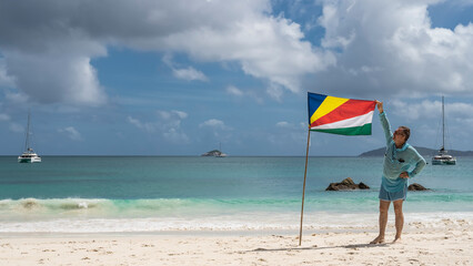 A man stands on a white sandy beach next to the flag of the Seychelles, holding a cloth around the...