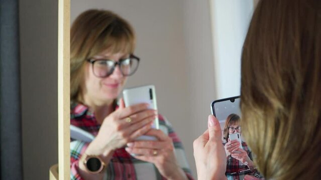 A girl with glasses takes a picture of herself on the phone in front of the mirror. Posing and taking selfies.