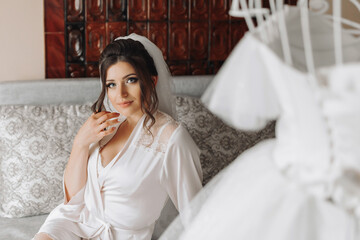 Obraz na płótnie Canvas A brunette bride in a dressing gown sits on a gray sofa, holding her black curls behind a mannequin with a wedding dress, against the background of painted walls. Wedding photo. Beautiful bride