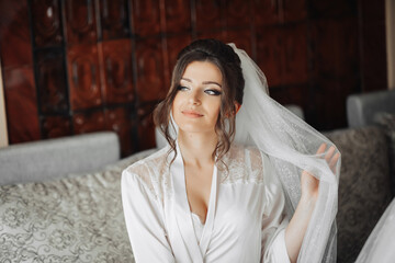 Portrait of the bride. A brunette bride is sitting on a gray sofa in a dressing gown, posing, holding her voluminous white veil. Gorgeous make-up and hair. Wedding photo. Beautiful bride