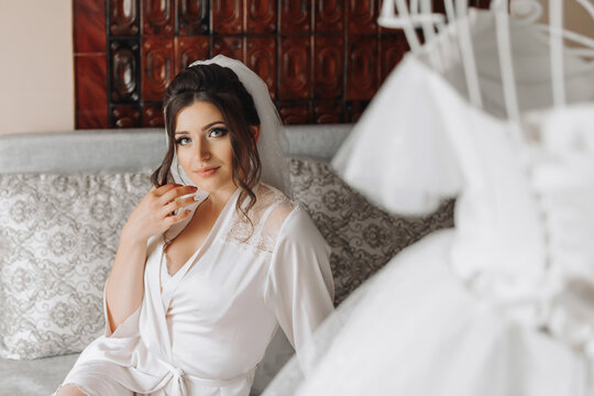 A brunette bride in a dressing gown sits on a gray sofa, holding her black curls behind a mannequin with a wedding dress, against the background of painted walls. Wedding photo. Beautiful bride