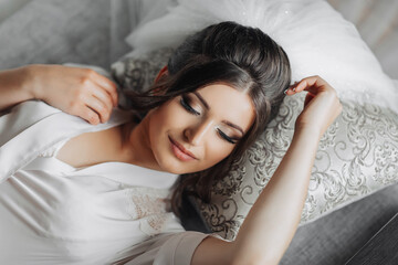 Brunette bride in a dressing gown posing for the camera while lying on a gray sofa. Portrait photo. Elegant hairstyle. Nice makeup. Beautiful hands. Morning of the bride.