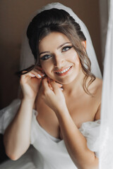A brunette bride in a white dress puts on her earrings and smiles sincerely at the camera. Open shoulders. Beautiful hands. Long veil. Morning of the bride.