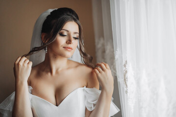 Fototapeta premium A brunette bride in a white dress holds her gorgeous curls and looks down. Open shoulders. Beautiful hands. Long veil. Morning of the bride.