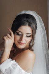 Fototapeta premium A brunette bride in a white dress holds her gorgeous curls and looks down. Open shoulders. Beautiful hands. Long veil. Morning of the bride.