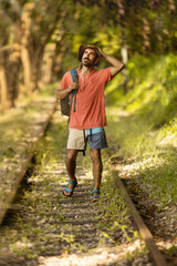 Naklejka na ściany i meble Backpacker man, looking off camera, walking along train tracks, observing Portugal's lush green forest in summertime. Wearing hat, pink t-shirt and short with sandals. Sunbeams.
