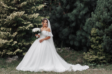 Obraz na płótnie Canvas The bride in a voluminous white dress and a long veil stands on a background of green conifers with a bouquet of white roses, holding her dress. Portrait of the bride. Beautiful makeup and hair.