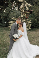 Portrait of the bride and groom in nature. The groom and the brunette bride in a white long dress are standing, kissing against the background of conifers. Stylish groom. Beautiful hair and makeup