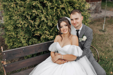 Portrait of the bride and groom in nature. A stylish groom and a brunette bride in a white long dress sit on a bench against a tree background, hug and sincerely smile. curly hair