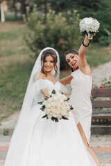 Wedding photo in nature. A brunette bride in a white long dress and her friend in a nude dress are...