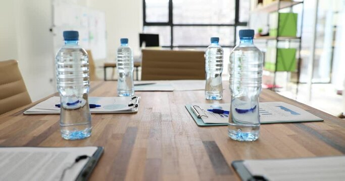 Bottles of water stand on wooden board table near clipboards with documents and pens. Spacious empty conference hall prepared for meeting in light office