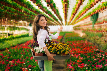 A smiling small business owner is standing in a greenhouse holding a crate of flowers.