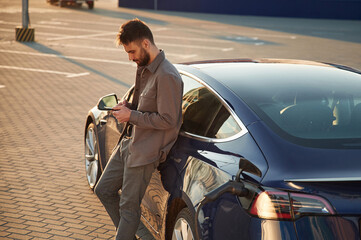 Leaning on the automobile and holding smartphone. On the charging station. Man is standing near his...