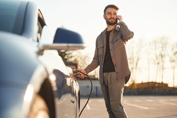 Business call by phone. Man is standing near his electric car outdoors