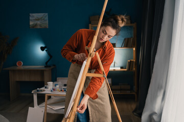 young beautiful brazilian woman artist in home studio puts canvas on easel and fixes it. Black hair, dressed in casual clothes. concept of creativity and hobby