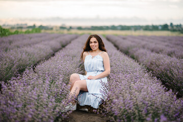 Fototapeta na wymiar A woman in a dress sitting in a lavender field at sunset. Female on a background of lavender flowers. France, Provence. Girl enjoy the floral glade, summer nature. Natural cosmetics and beauty concept